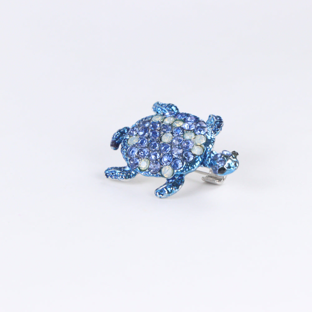 Metallic blue sea turtle brooch pin encrusted with sapphire blue and opal-tone crystals on a white background by Delemon