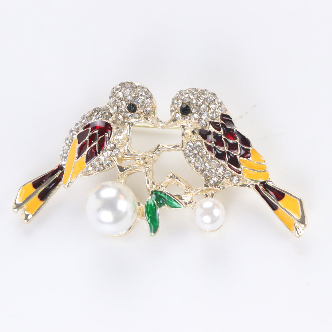 Gold-tone lovebirds brooch pin with clear crystal paved heads, maroon and yellow enamel wings, green leaves, and hanging pearls on a white background by Delemon