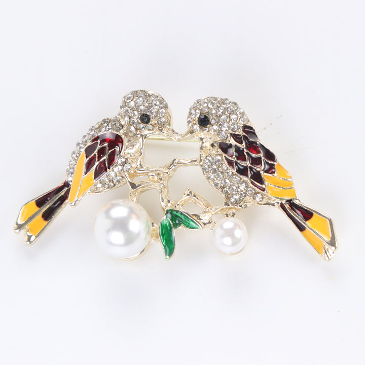 Gold-tone lovebirds brooch pin with clear crystal paved heads, maroon and yellow enamel wings, green leaves, and hanging pearls on a white background by Delemon