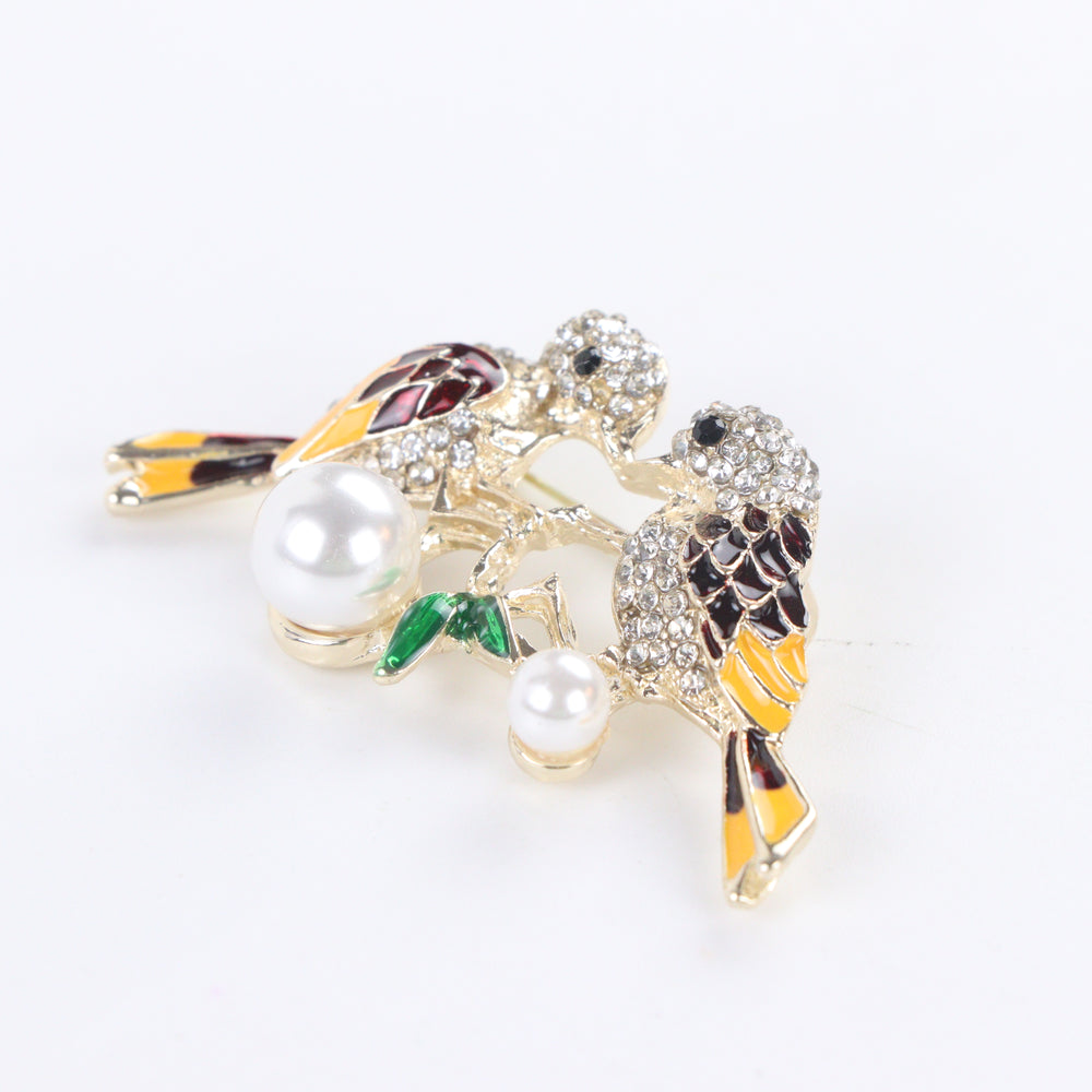 Gold-tone lovebirds brooch pin with clear crystal paved heads, maroon and yellow enamel wings, green leaves, and hanging pearls on a white background by Delemon