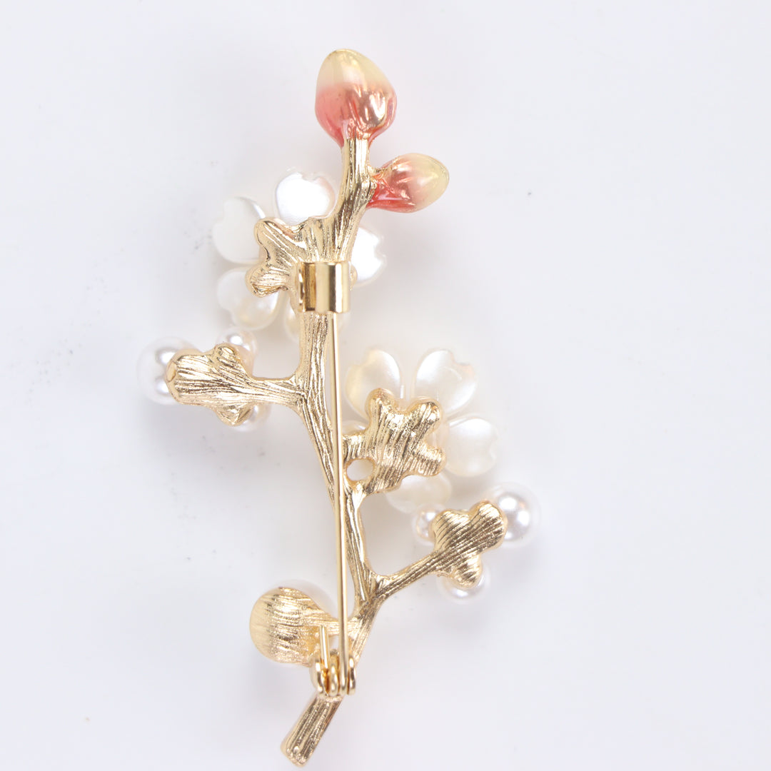 Gold-tone cherry blossom branch brooch pin with pearlescent white enamel flowers, ruby pink crystals, and pearl clusters on a white background by Delemon