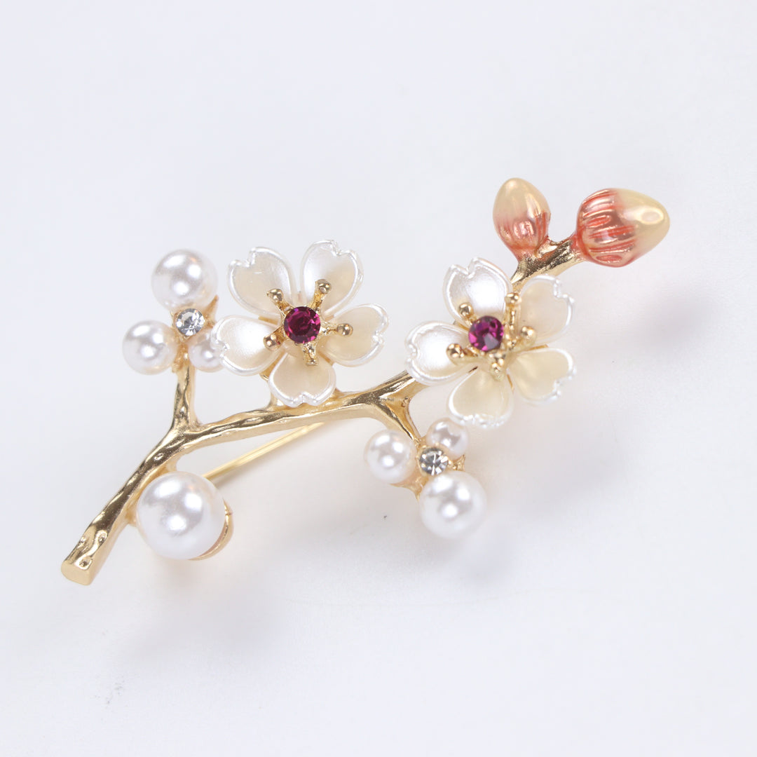 Gold-tone cherry blossom branch brooch pin with pearlescent white enamel flowers, ruby pink crystals, and pearl clusters on a white background by Delemon