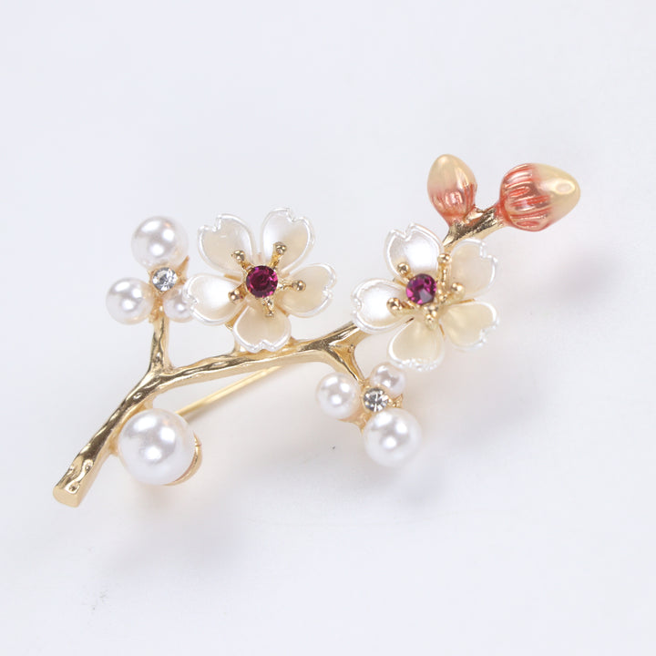 Gold-tone cherry blossom branch brooch pin with pearlescent white enamel flowers, ruby pink crystals, and pearl clusters on a white background by Delemon