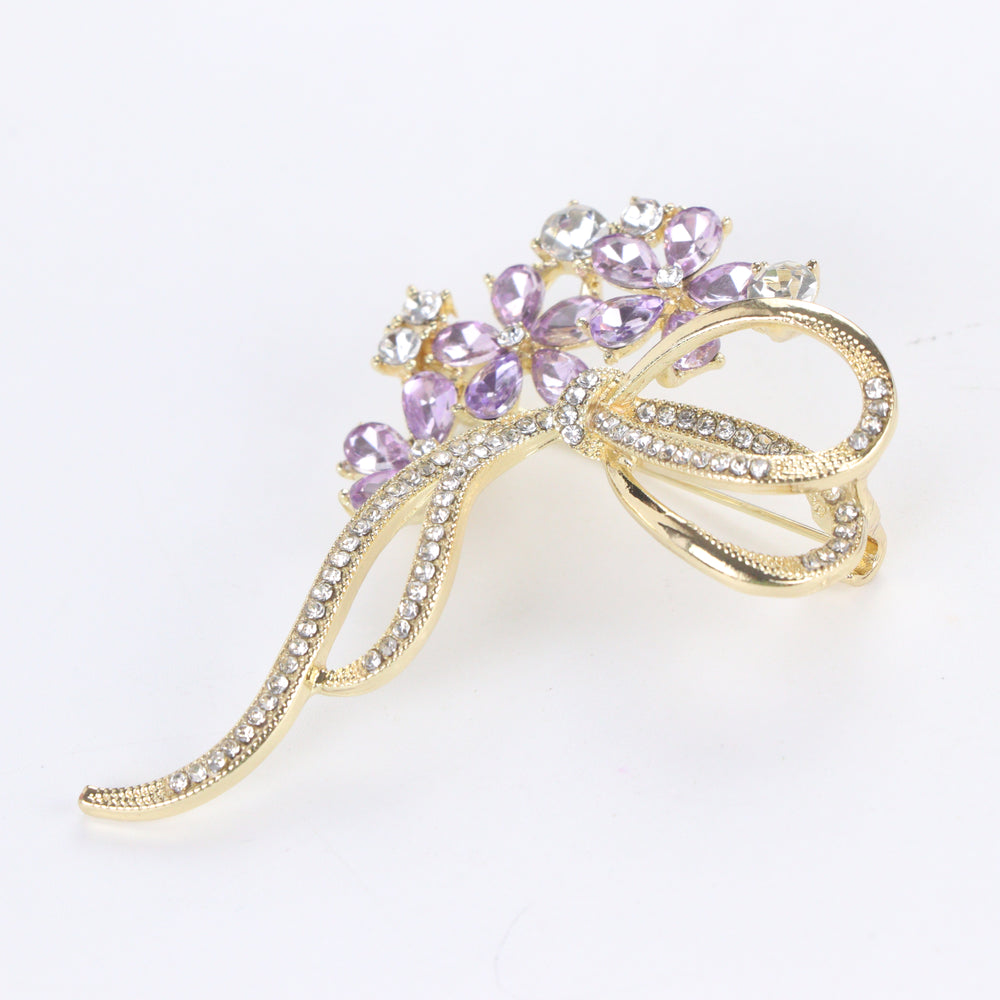 Gold-tone floral bow brooch pin featuring purple tear-drop crystal blossoms and clear pavé rhinestone ribbons on a white background by Delemon