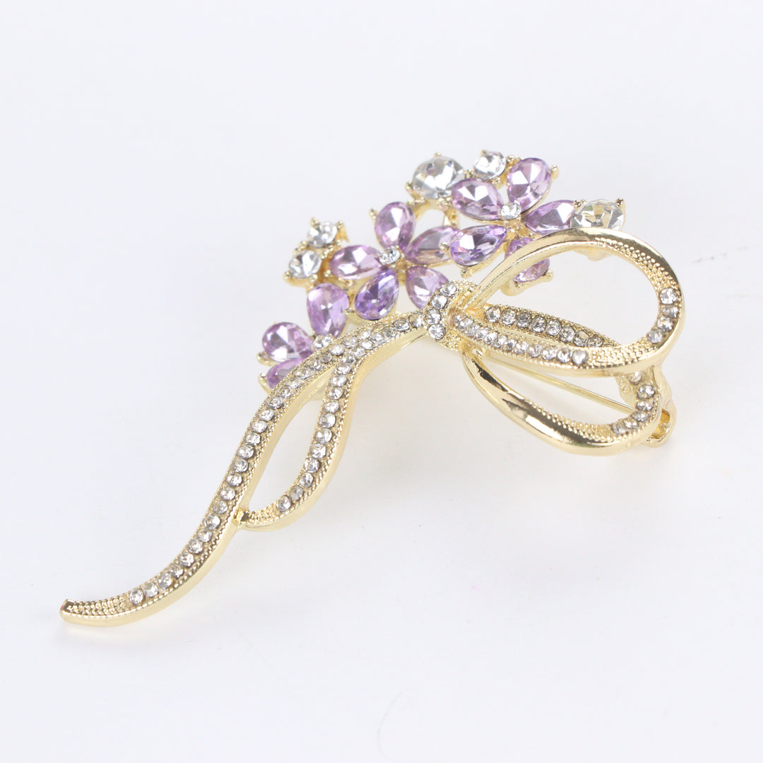 Gold-tone floral bow brooch pin featuring purple tear-drop crystal blossoms and clear pavé rhinestone ribbons on a white background by Delemon