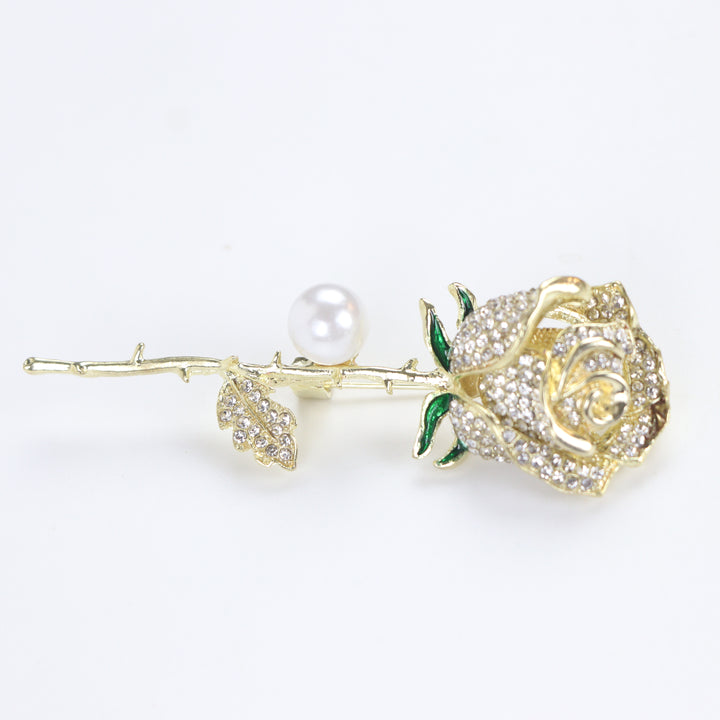 Gold rose brooch pin encrusted with pave crystals, green enamel accents, and a pearl on a white background by Delemon