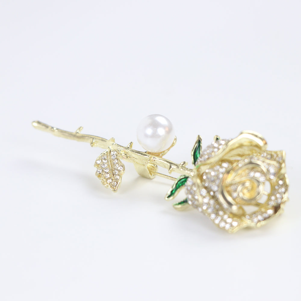 Gold rose brooch pin encrusted with pave crystals, green enamel accents, and a pearl on a white background by Delemon