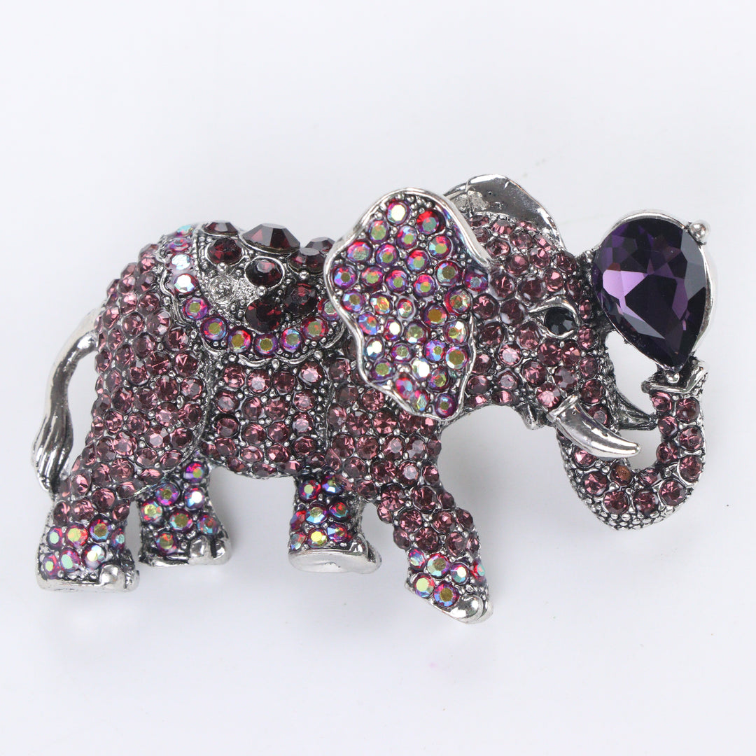 Antique silver-tone elephant brooch pin pavé-set with purple and iridescent crystals holding a pear-cut purple jewel in its raised trunk on a white background by Delemon