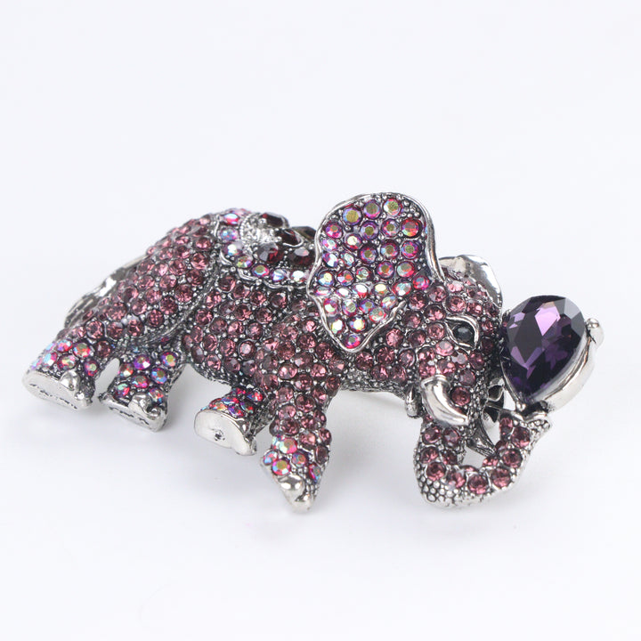 Antique silver-tone elephant brooch pin pavé-set with purple and iridescent crystals holding a pear-cut purple jewel in its raised trunk on a white background by Delemon