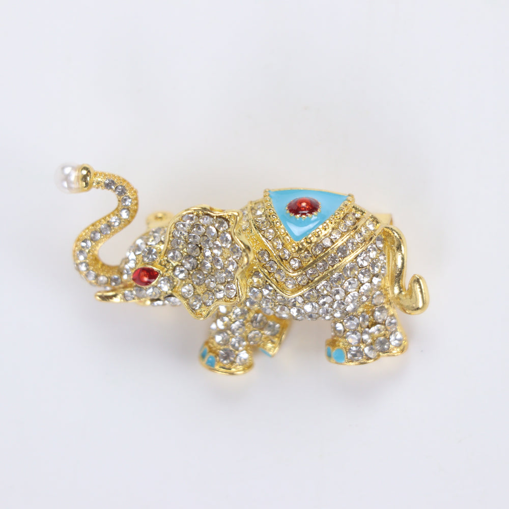 Sparkling gold elephant brooch pin with paved crystals, turquoise enamel saddle, and a pearl trunk tip on a white background by Delemon