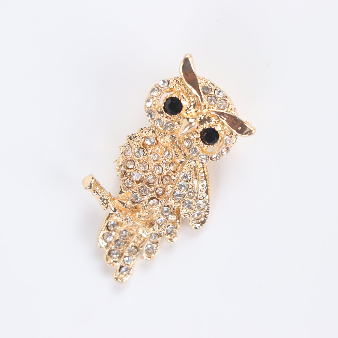 Gold owl brooch pin encrusted with clear crystals and black crystal eyes perched on a branch against a white background by Delemon