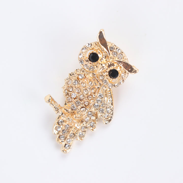 Gold owl brooch pin encrusted with clear crystals and black crystal eyes perched on a branch against a white background by Delemon