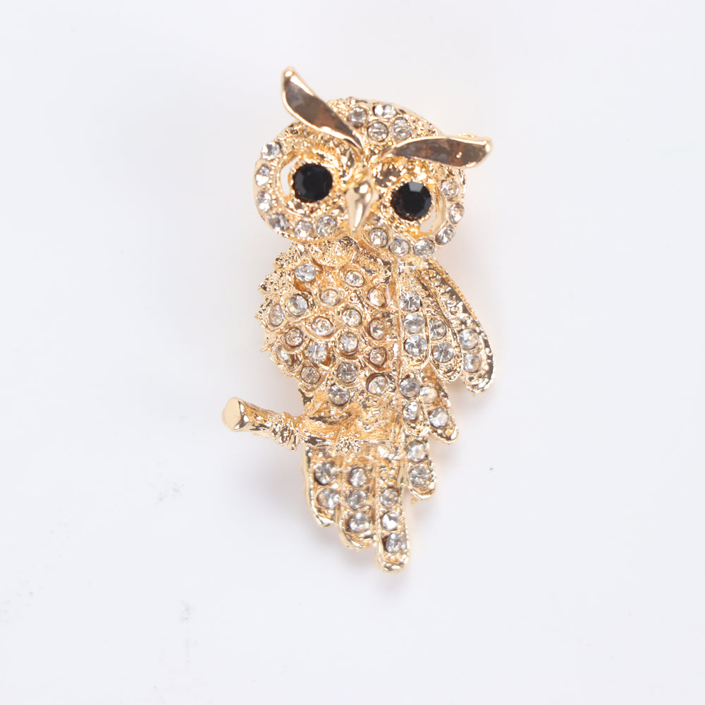 Gold owl brooch pin encrusted with clear crystals and black crystal eyes perched on a branch against a white background by Delemon