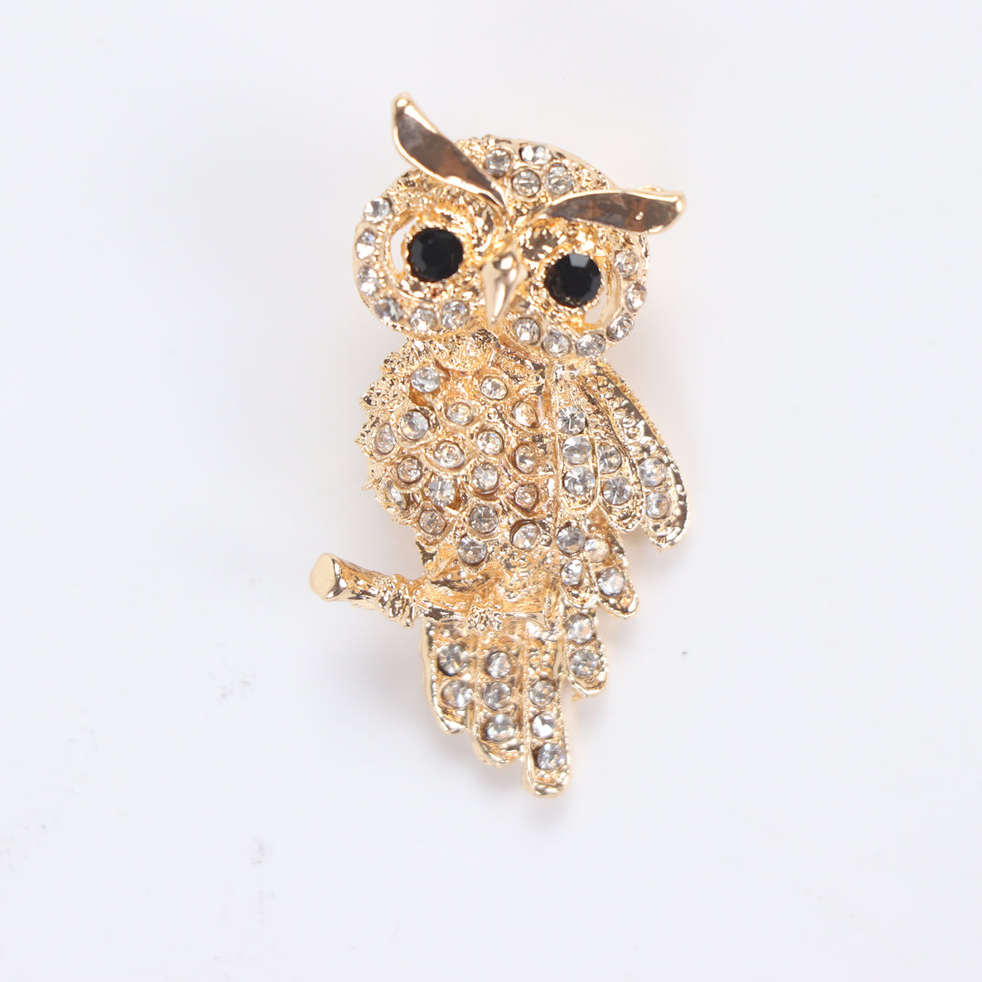 Gold owl brooch pin encrusted with clear crystals and black crystal eyes perched on a branch against a white background by Delemon