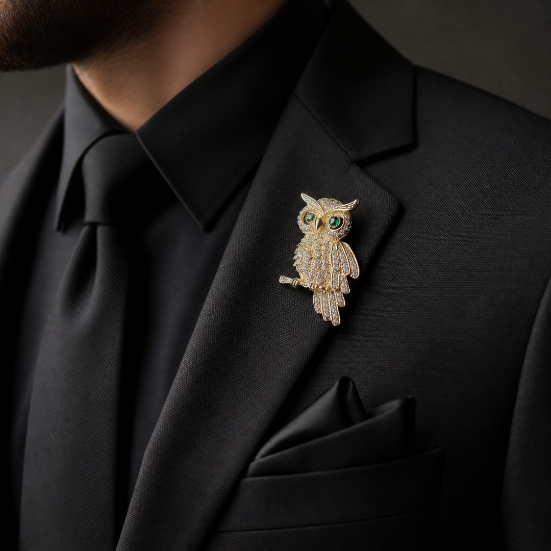 Gold owl brooch pin encrusted with clear crystals and green crystal eyes perched on a branch against a white background by Delemon