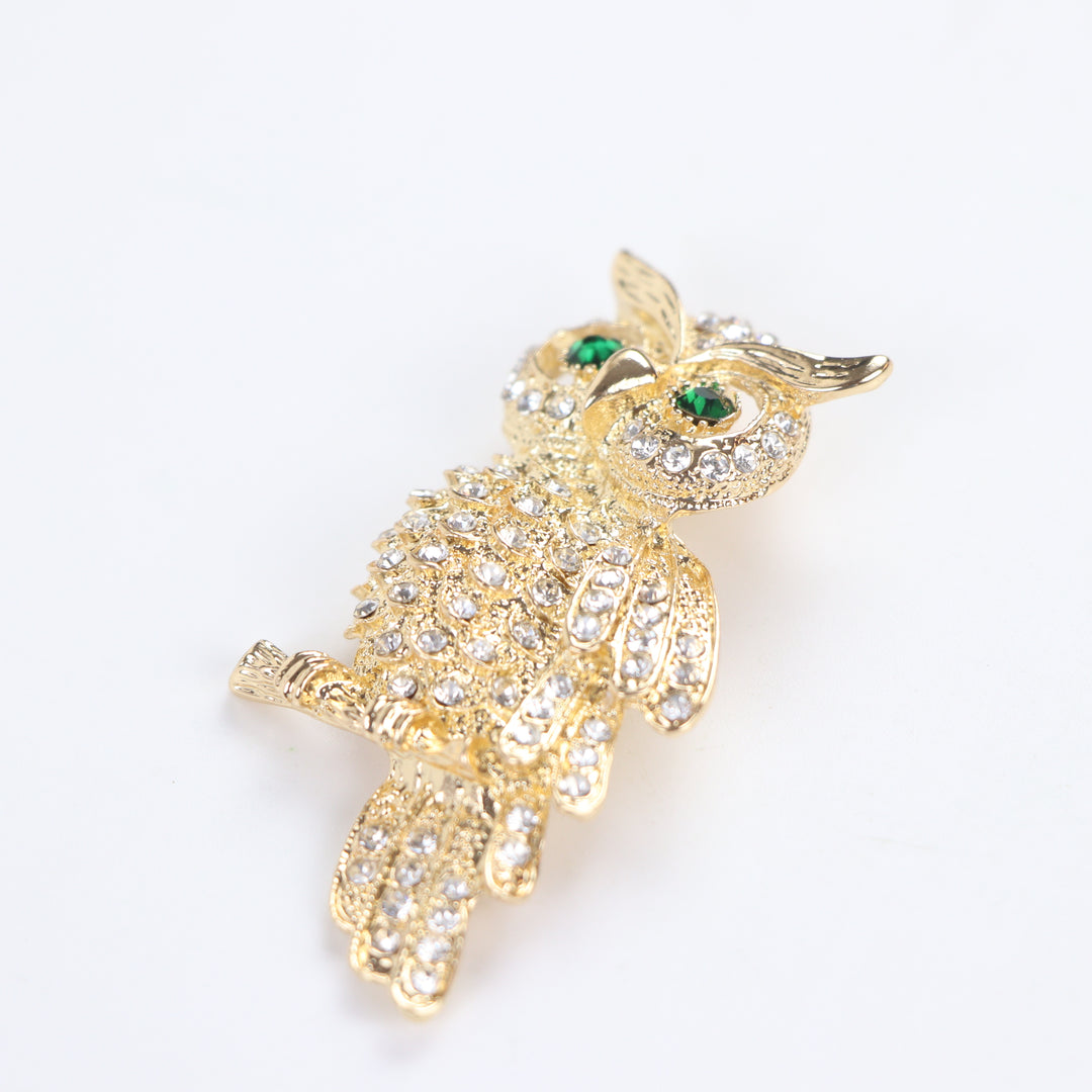 Gold owl brooch pin encrusted with clear crystals and green crystal eyes perched on a branch against a white background by Delemon
