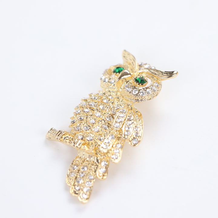 Gold owl brooch pin encrusted with clear crystals and green crystal eyes perched on a branch against a white background by Delemon