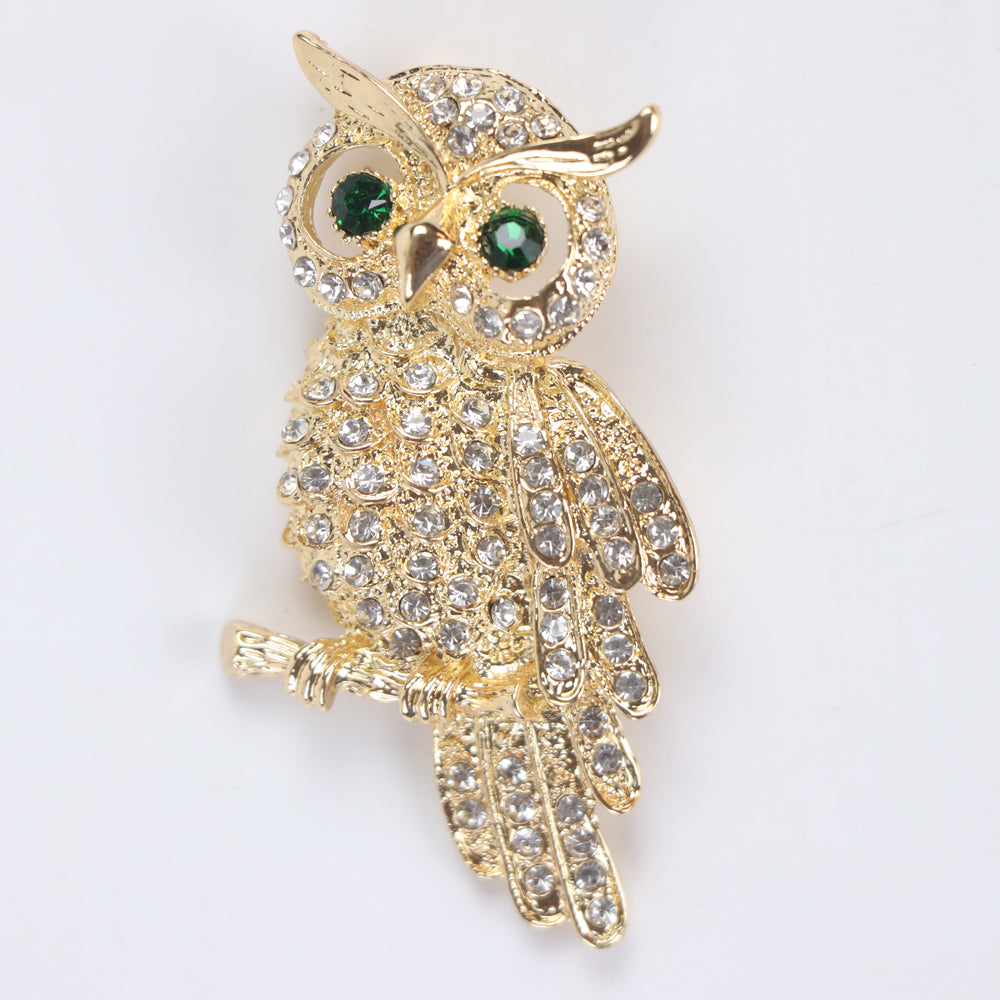 Gold owl brooch pin encrusted with clear crystals and green crystal eyes perched on a branch against a white background by Delemon