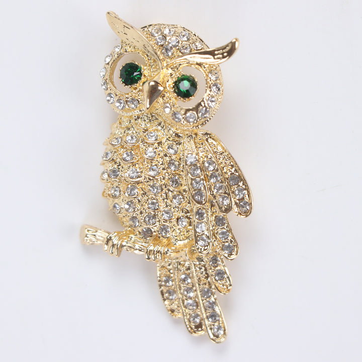 Gold owl brooch pin encrusted with clear crystals and green crystal eyes perched on a branch against a white background by Delemon