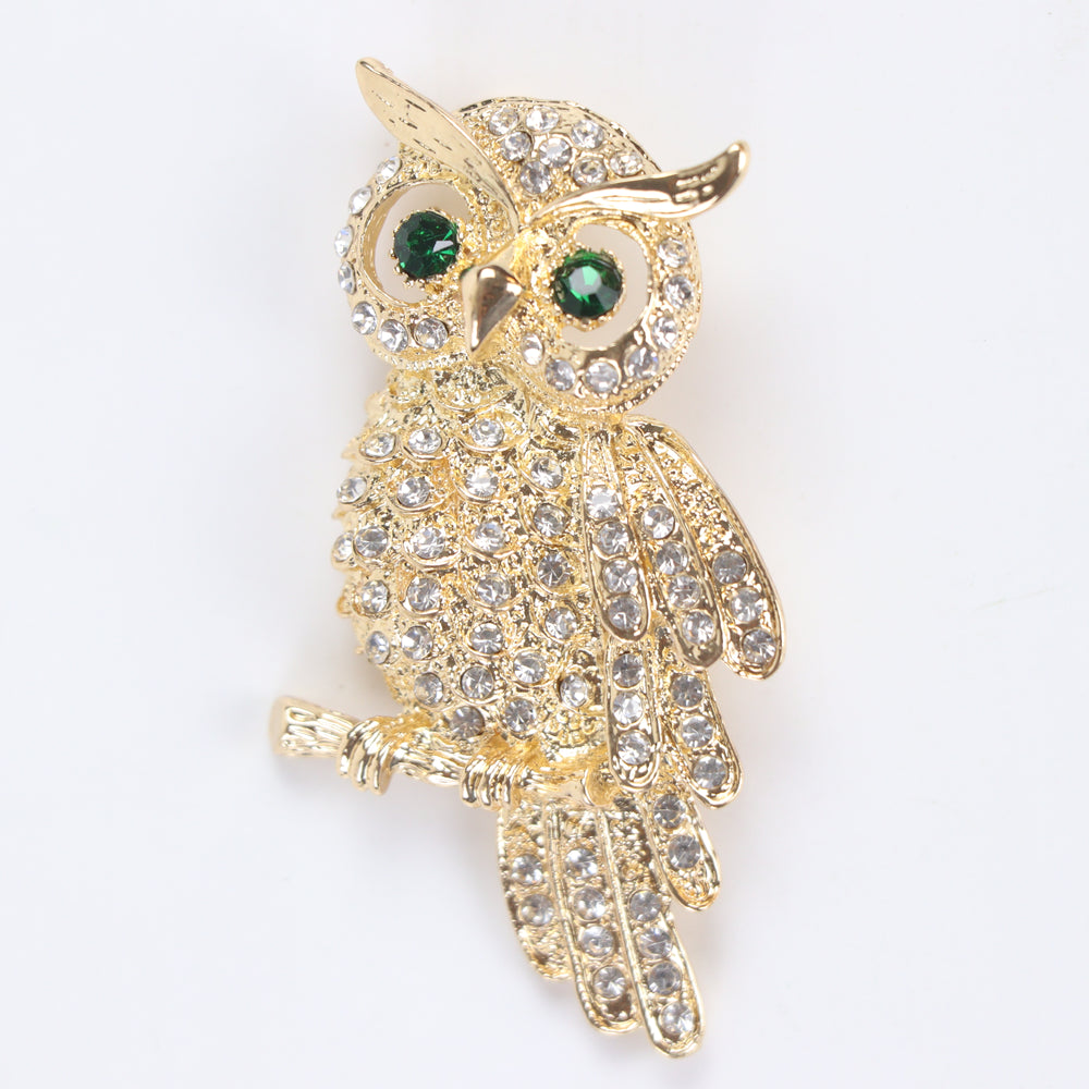 Gold owl brooch pin encrusted with clear crystals and green crystal eyes perched on a branch against a white background by Delemon