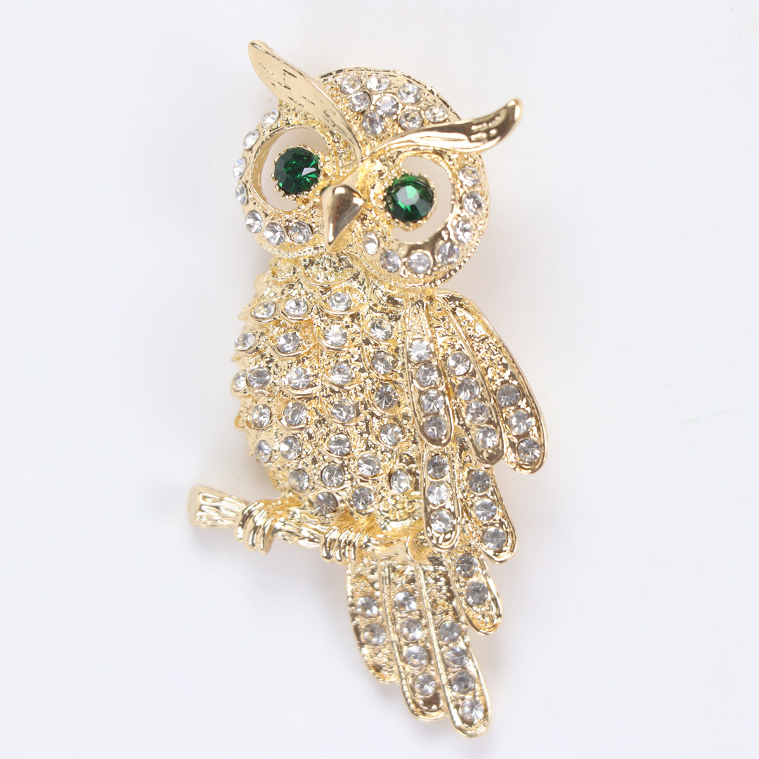 Gold owl brooch pin encrusted with clear crystals and green crystal eyes perched on a branch against a white background by Delemon
