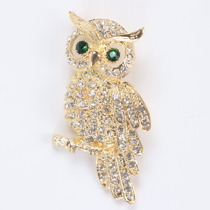 Gold owl brooch pin encrusted with clear crystals and green crystal eyes perched on a branch against a white background by Delemon