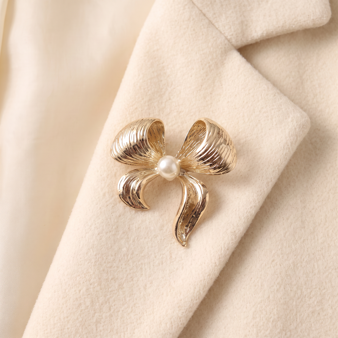 Textured gold ribbon bow brooch pin featuring a centered pearl accent against a white background by Delemon
