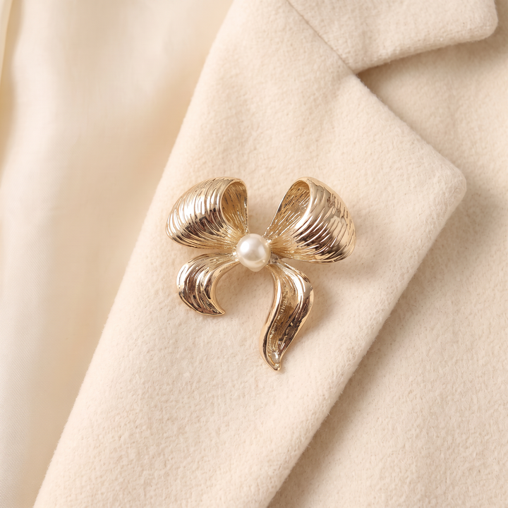 Textured gold ribbon bow brooch pin featuring a centered pearl accent against a white background by Delemon