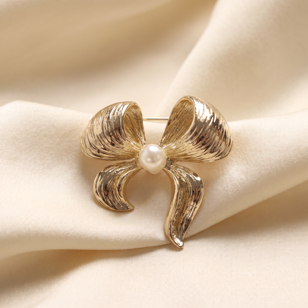 Textured gold ribbon bow brooch pin featuring a centered pearl accent against a white background by Delemon