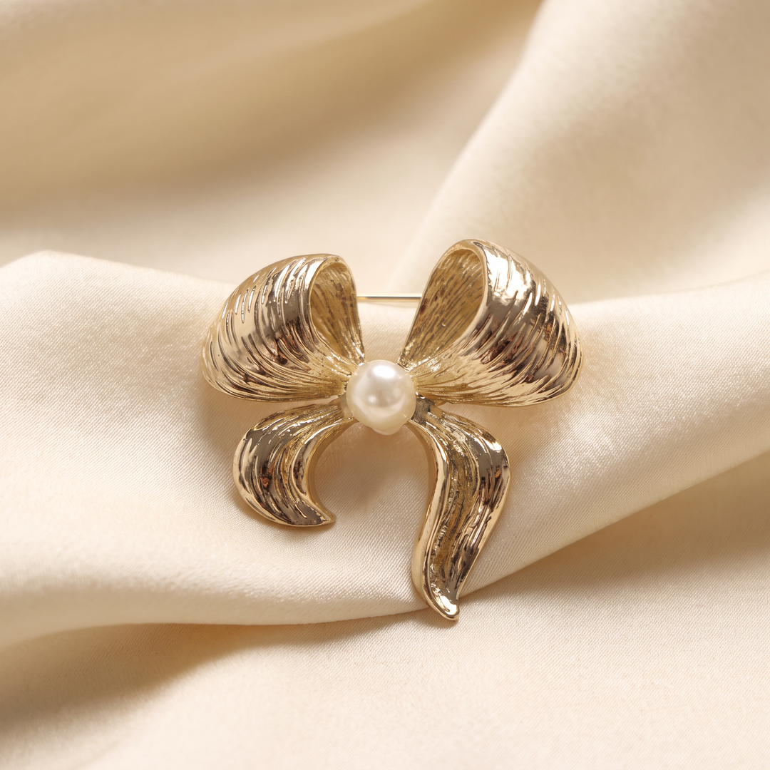 Textured gold ribbon bow brooch pin featuring a centered pearl accent against a white background by Delemon