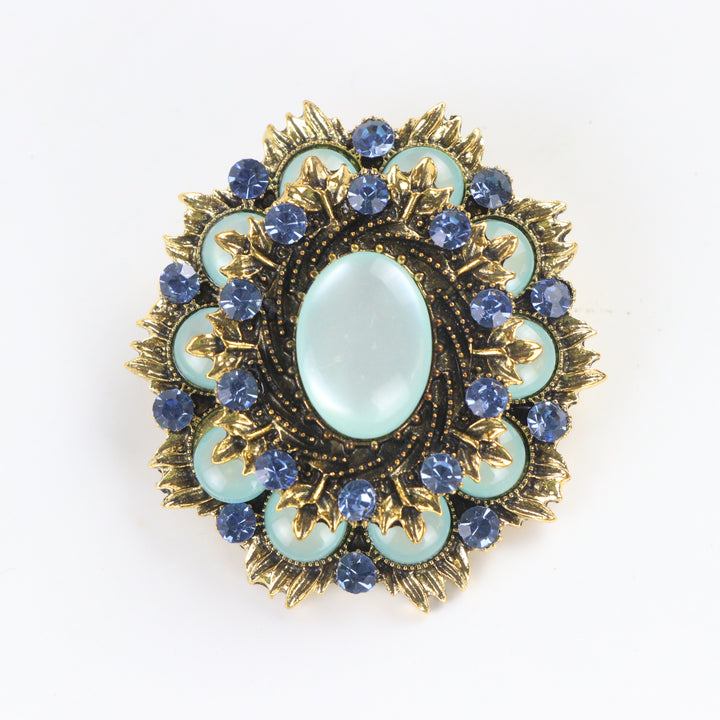 Ornate antique gold-tone round medallion brooch pin featuring a mint green cabochon center and sapphire blue crystal halo on a white background by Delemon