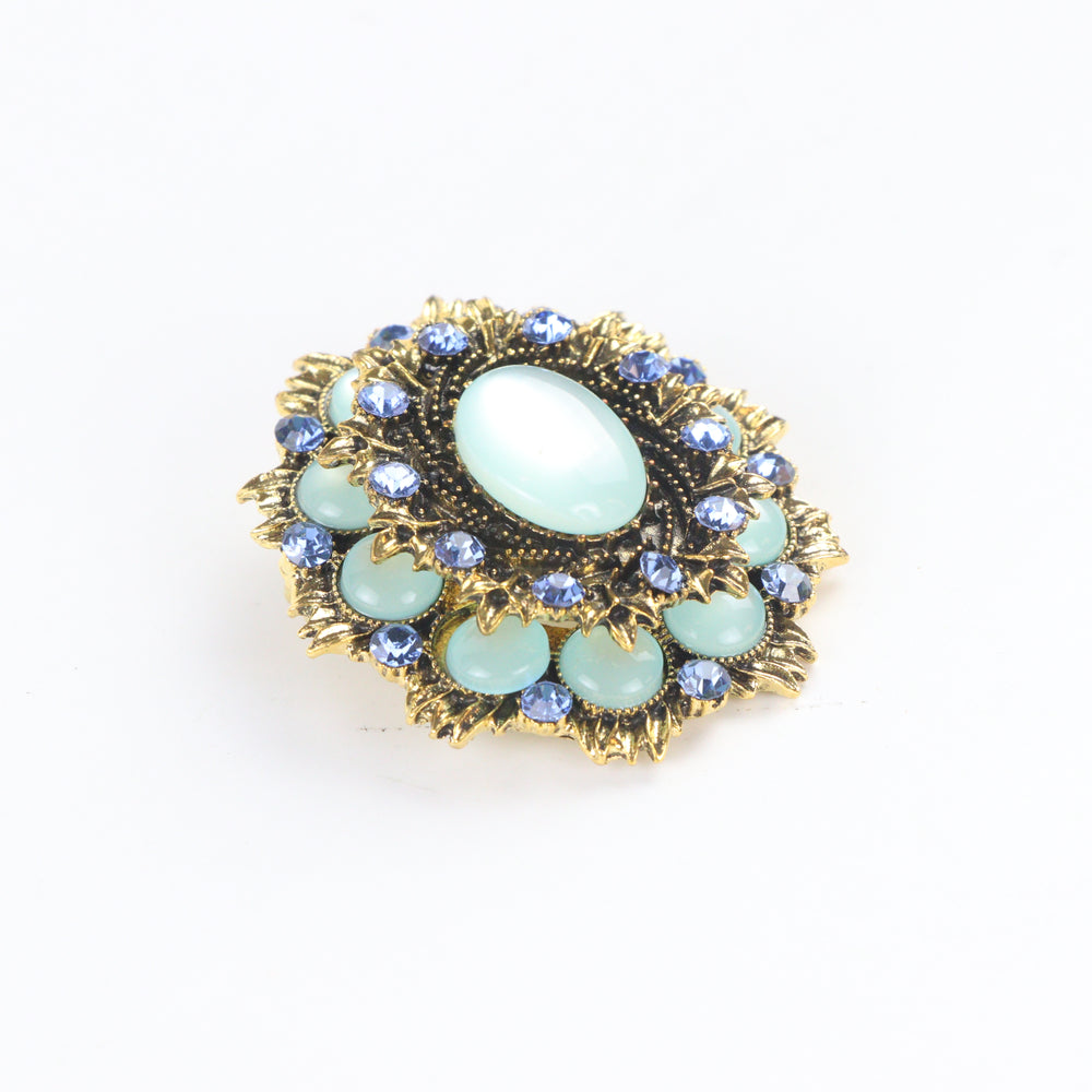 Ornate antique gold-tone round medallion brooch pin featuring a mint green cabochon center and sapphire blue crystal halo on a white background by Delemon