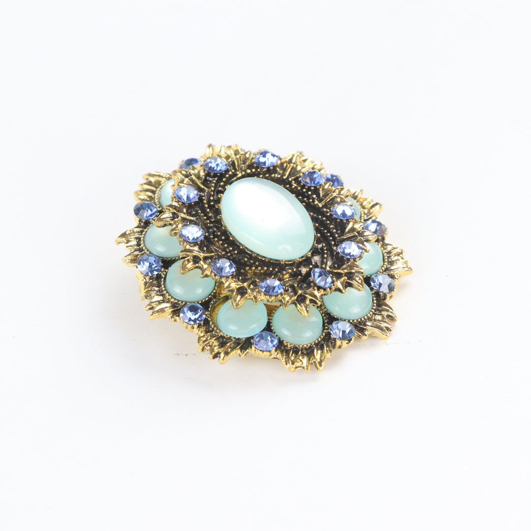 Ornate antique gold-tone round medallion brooch pin featuring a mint green cabochon center and sapphire blue crystal halo on a white background by Delemon